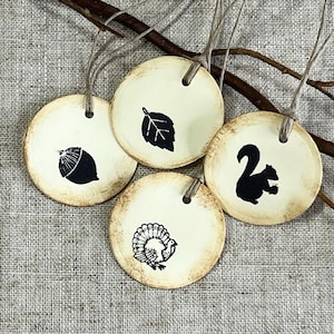 Four round cream gift tags with hand-distressed edges displayed on natural linen with rustic branches. The hand-stamped black designs feature an acorn, leaf, squirrel and turkey, with each tag finished with cord.