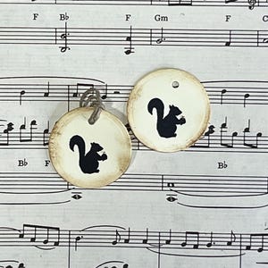 Two round cream gift tags with black hand-stamped squirrel designs displayed against vintage sheet music. The tags feature distressed edges, a subtle raised border and cord ties.