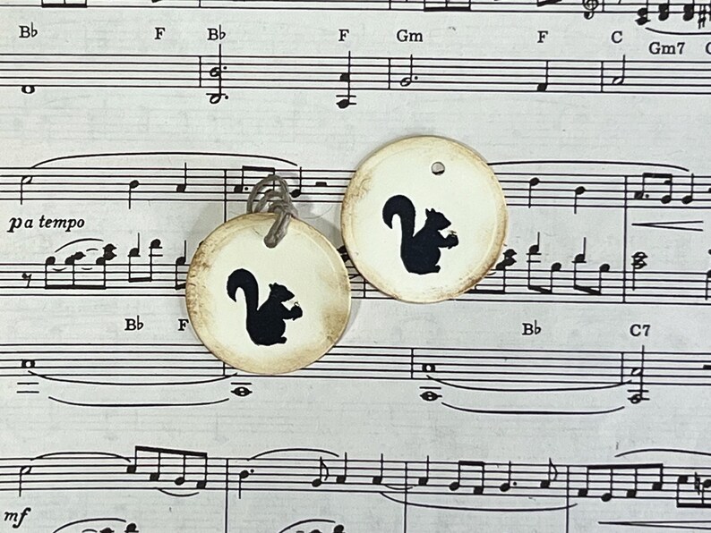 Two round cream gift tags with black hand-stamped squirrel designs displayed against vintage sheet music. The tags feature distressed edges, a subtle raised border and cord ties.