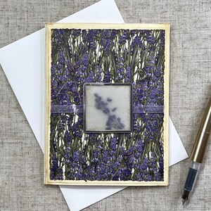 May include: A handmade greeting card with a lavender floral design. The card features a square, framed image of lavender and a decorative ribbon. A gold fountain pen rests on the surface next to the card and a white envelope.