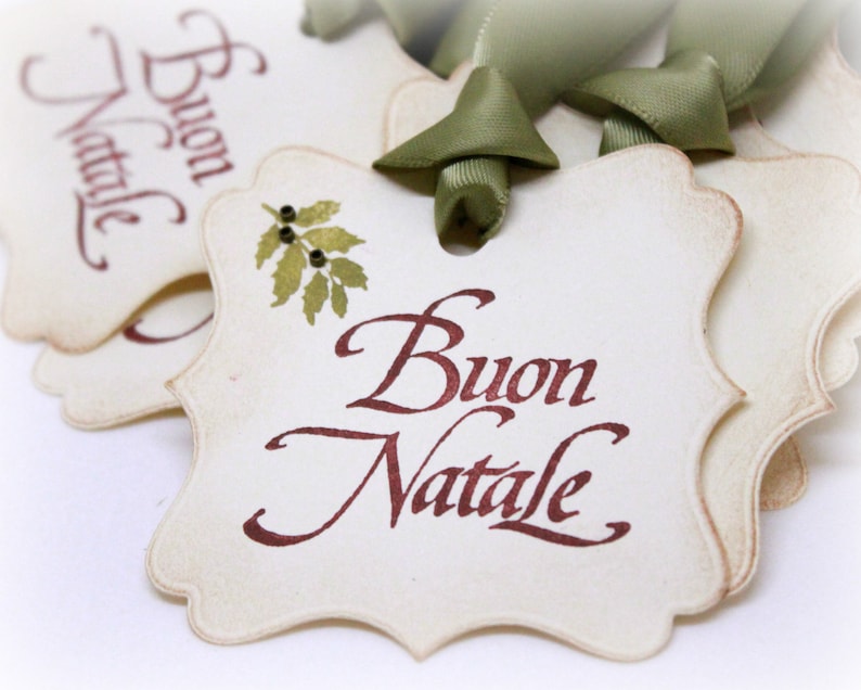 May include: Two cream-colored paper gift tags with scalloped edges. The tags are tied together with a green ribbon. The top tag has the words 'Buon Natale' written in brown ink. The bottom tag has a small green leaf design and the words 'Buon Natale' written in brown ink.