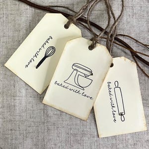 Op de afbeelding: Drie crèmekleurige cadeaulabels met zwarte lijntekeningen van bakgereedschap: een garde, een keukenmixer en een deegroller. Elk label heeft de zin "baked with love" in een handschrift. De labels zijn vastgebonden met touw.