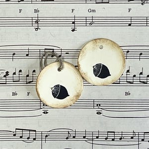 Two round cream gift tags with hand-stamped black acorn designs displayed on vintage sheet music. The tags have hand-distressed edges, a subtle raised border and cord ties.