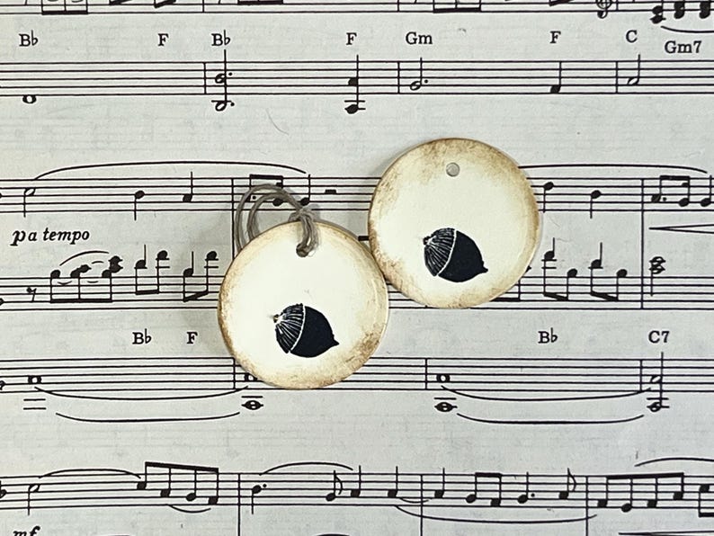 Two round cream gift tags with hand-stamped black acorn designs displayed on vintage sheet music. The tags have hand-distressed edges, a subtle raised border and cord ties.