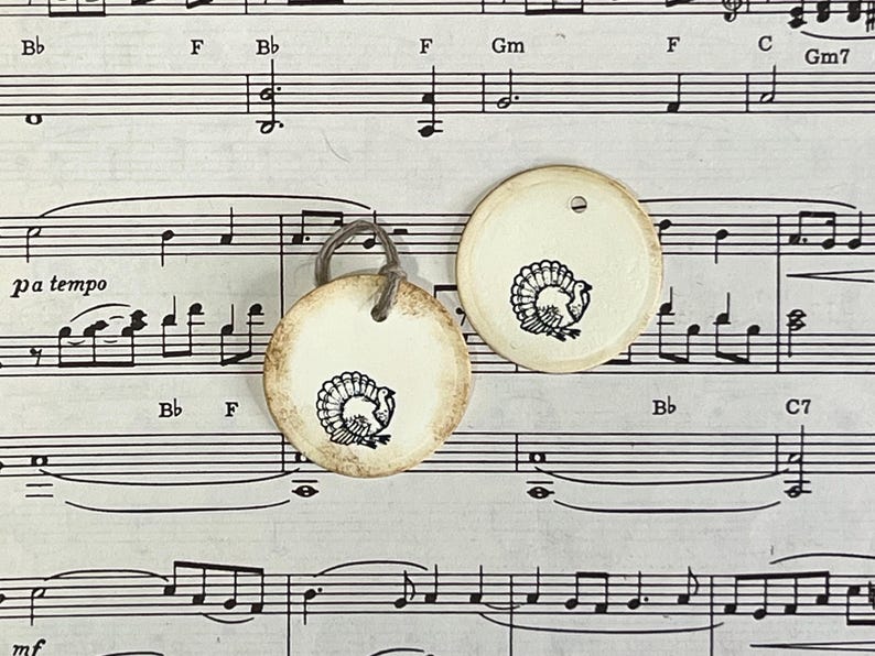 Two round cream gift tags featuring hand-stamped black turkey designs on vintage sheet music. Each tag has softly distressed edges, a subtle raised border and a cord tie.