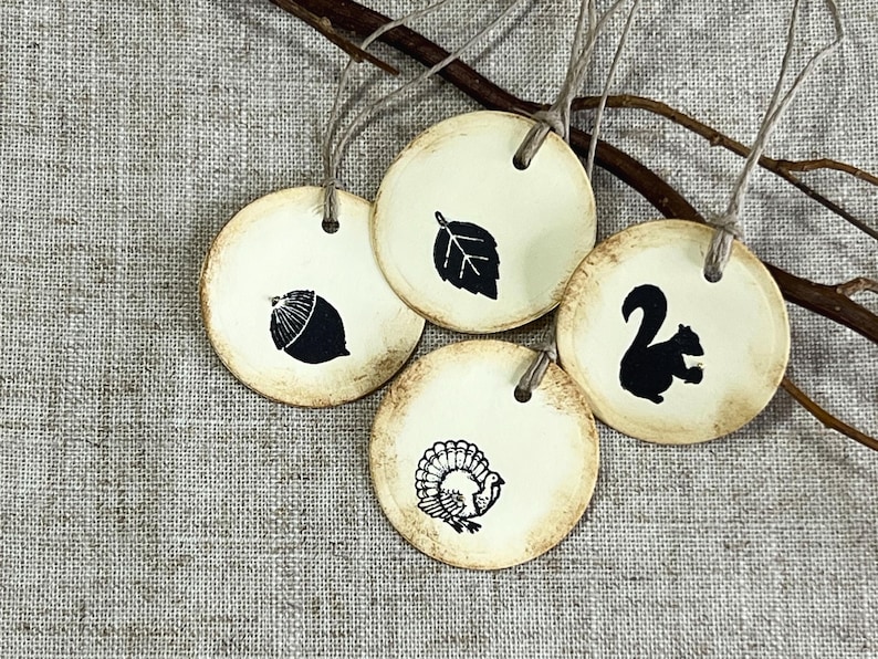 Four round cream gift tags with hand-distressed edges displayed on natural linen with rustic branches. The hand-stamped black designs feature an acorn, leaf, squirrel and turkey, with each tag finished with cord.