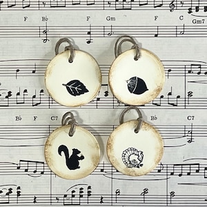 Four round hand-stamped gift tags arranged on vintage sheet music, showing the Forest Life acorn, leaf, squirrel and turkey designs. The cream tags have distressed edges, a subtle raised border and cord ties.
