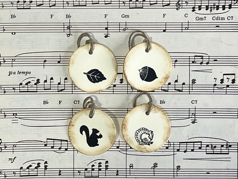 Four round hand-stamped gift tags arranged on vintage sheet music, showing the Forest Life acorn, leaf, squirrel and turkey designs. The cream tags have distressed edges, a subtle raised border and cord ties.