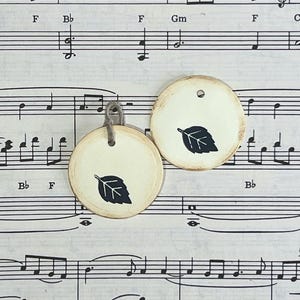 Two round cream gift tags with black hand-stamped leaf designs displayed on vintage sheet music. The tags have softly distressed edges, and one tag is finished with a gray cord tie.