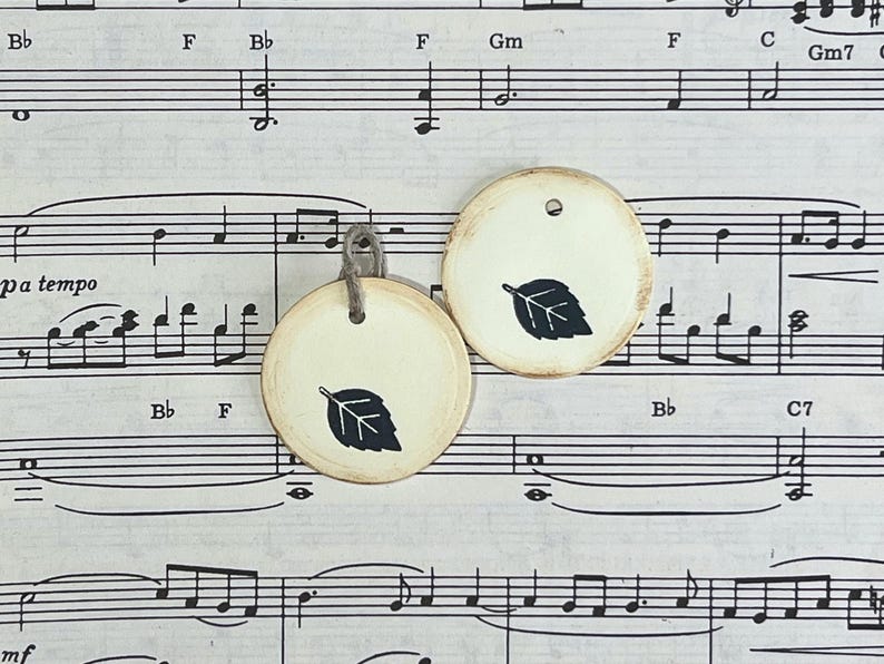 Two round cream gift tags with black hand-stamped leaf designs displayed on vintage sheet music. The tags have softly distressed edges, and one tag is finished with a gray cord tie.