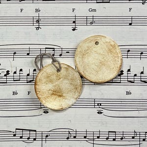 Reverse views of a round cream gift tag displayed on vintage sheet music. The reverse is left blank with hand-distressed aging across the surface and edges, while the second tag shows the matching distressed finish.