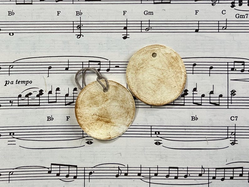 Reverse views of a round cream gift tag displayed on vintage sheet music. The reverse is left blank with hand-distressed aging across the surface and edges, while the second tag shows the matching distressed finish.