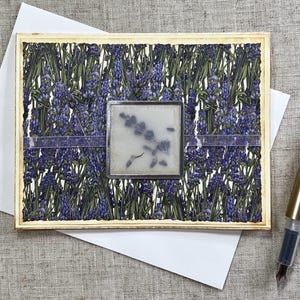 May include: A handmade greeting card with a floral design featuring purple flowers and green leaves. The card has a wooden frame and a clear, square embellishment. A white envelope and a gold fountain pen are also present.