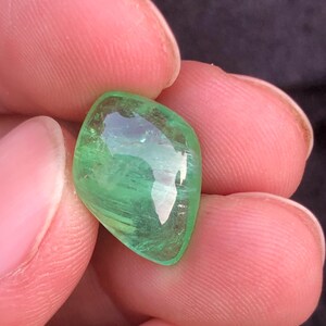 Op de afbeelding: Een levendige, smaragdgroene edelsteen met een gepolijste, afgeronde vorm. De edelsteen wordt tussen vingers gehouden, wat de helderheid en interne structuur laat zien. De kleur van de steen is een rijk, verzadigd groen, dat licht reflecteert en een sprankelend effect creëert.