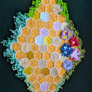 May include: A handmade crochet piece featuring a honeycomb pattern in shades of yellow, tan, and white. The edges are trimmed with green fringe, and several colorful crochet flowers are attached, including red, blue, and purple.