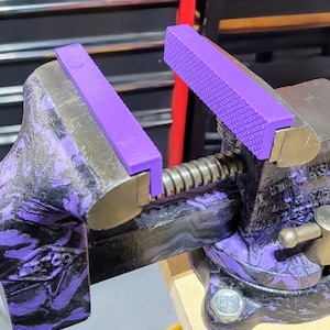 May include: A purple and black marbled vise with purple jaw protectors. The vise is open, revealing the screw mechanism. The jaw protectors have a textured surface. The vise is on a wooden surface.