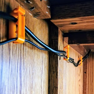 May include: Orange cable clips attached to a wooden surface, holding black electrical cords in place.