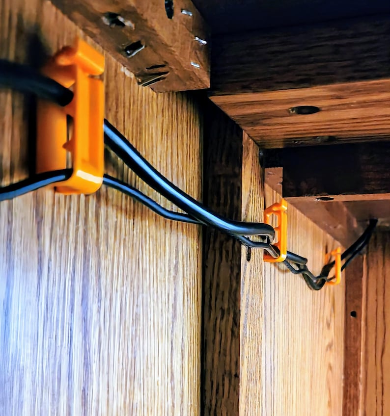 May include: Orange cable clips attached to a wooden surface, holding black electrical cords in place.