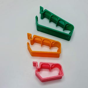 May include: Three plastic cable clips in different sizes. The largest clip is green, the middle clip is orange, and the smallest clip is pink.