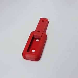 May include: A bright red plastic bracket with a rectangular base and a hinged, elongated arm. The base features two circular holes, and the arm has two additional holes. The bracket is designed for mounting or securing objects.