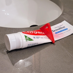 Puede incluir: Un dispensador de pasta de dientes de plástico rojo que sostiene un tubo de pasta de dientes Colgate Sensitive con el texto "MAXIMUM STRENGTH" y "COMPLETE PROTECTION" en el tubo.