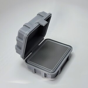 May include: A gray, open, rectangular case with a hinged lid. The interior features a ridged, dark gray surface. The case is made of a durable material and has a secure latching mechanism, suggesting it is designed for protection.