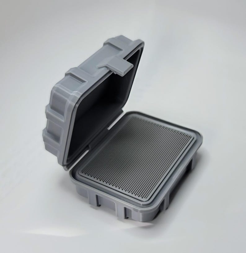 May include: A gray, open, rectangular case with a hinged lid. The interior features a ridged, dark gray surface. The case is made of a durable material and has a secure latching mechanism, suggesting it is designed for protection.
