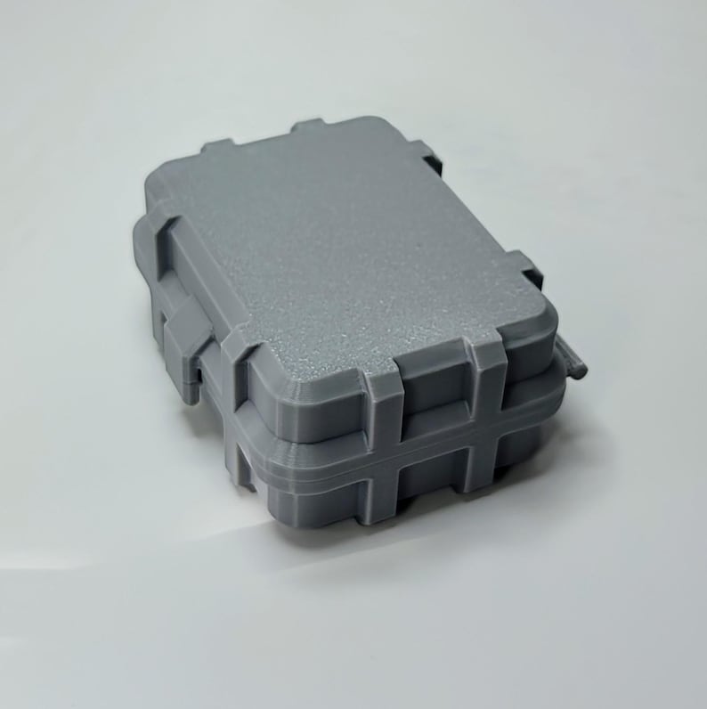 May include: A gray, rectangular, small storage case with a textured surface. The case has a hinged lid and a secure latching mechanism. The case is designed to protect its contents and is suitable for various applications.