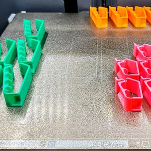 May include: A close-up of a 3D printed object on a textured surface. The object is made up of multiple pieces in three different colors: green, orange, and pink.