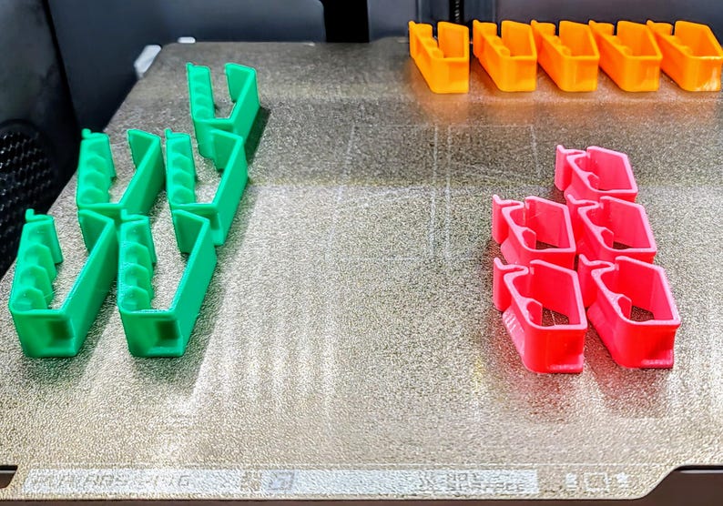 May include: A close-up of a 3D printed object on a textured surface. The object is made up of multiple pieces in three different colors: green, orange, and pink.