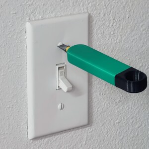 May include: A green and black tool is being used to remove a light switch cover from a white wall. The switch is in the "on" position.