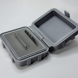 May include: Open gray plastic container with a hinged lid. The interior features a slotted tray holding several metal blades. The container is designed to protect and store sharp objects, such as razor blades or craft knives.