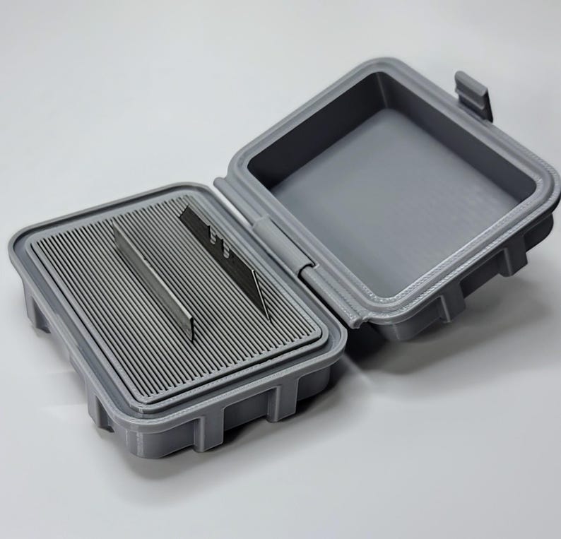 May include: Open gray plastic container with a hinged lid. The interior features a slotted tray holding several metal blades. The container is designed to protect and store sharp objects, such as razor blades or craft knives.