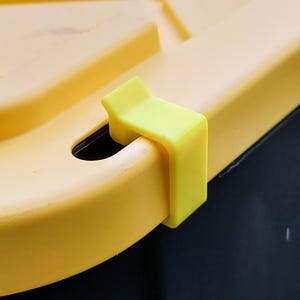 May include: Close-up of a yellow plastic component with a right-angled, clip-like design. The clip is attached to a curved yellow surface, with a dark background. The component appears to be part of a larger structure.