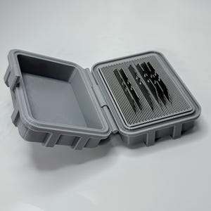 May include: Open gray plastic case with a hinged lid. The case is rectangular with a textured surface and a latch. Inside, there are several dark blades arranged in a slotted tray. The case is designed for storage and organization.