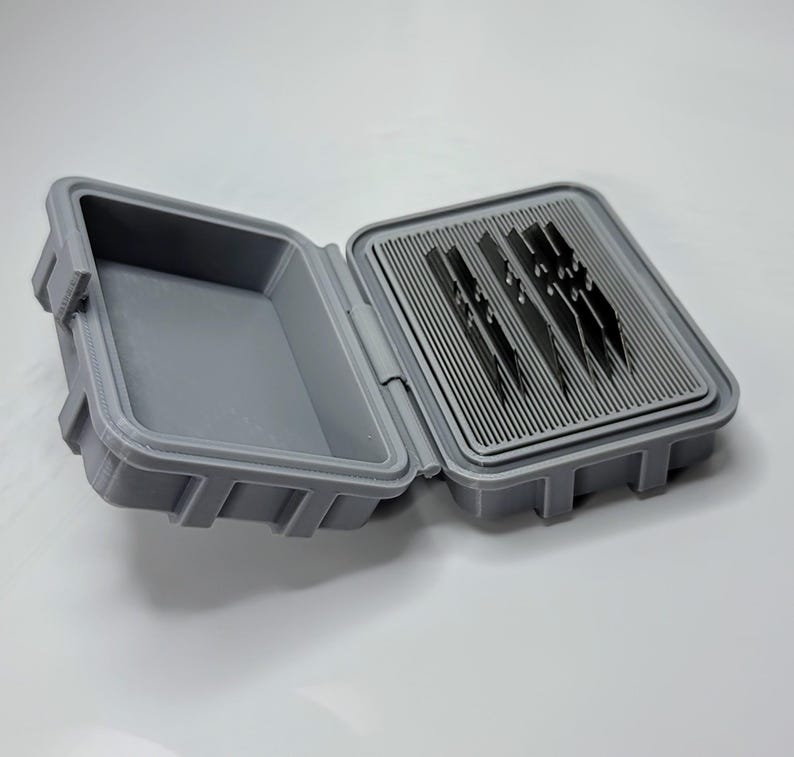 May include: Open gray plastic case with a hinged lid. The case is rectangular with a textured surface and a latch. Inside, there are several dark blades arranged in a slotted tray. The case is designed for storage and organization.