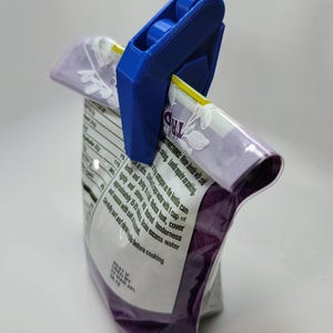 May include: A blue plastic bag clip with a lever and two wheels. The clip is holding a purple and white bag with text that reads "Rinse lentils before cooking. Lentils require no soaking.  The lentils can be eaten in the same size as the bag.  Bring to a boil with 1 cup of water and simmer until heated, cover tightly and allow to stand with heat off until desired tenderness. Drain excess water and season with salt and fresh herbs.  Carefully sort and rinse before cooking.  Best if used by 03 Mar 20L 08.42"