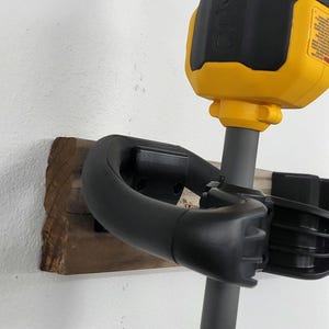 May include: A black and yellow electric weed trimmer with a black handle and a gray shaft is mounted on a wooden wall mount.