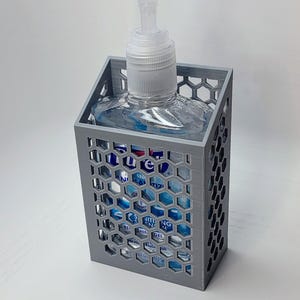 May include: A gray 3D printed holder with a honeycomb design holds a clear bottle of hand sanitizer with a pump dispenser. The bottle label reads "Purell" and "Antibacterial Hand Sanitizer".