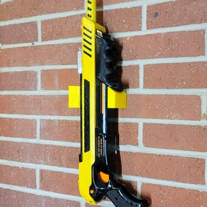 May include: A yellow and black toy blaster gun with an orange tip is mounted on a brick wall. The gun has a black handle and the text "DO NOT SHOOT IN FACE OR EYES!" is visible. The blaster is held in place by a yellow bracket.