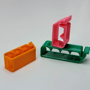 May include: Three 3D printed plastic cable clips in orange, green, and pink. The clips are designed to hold cables in place.