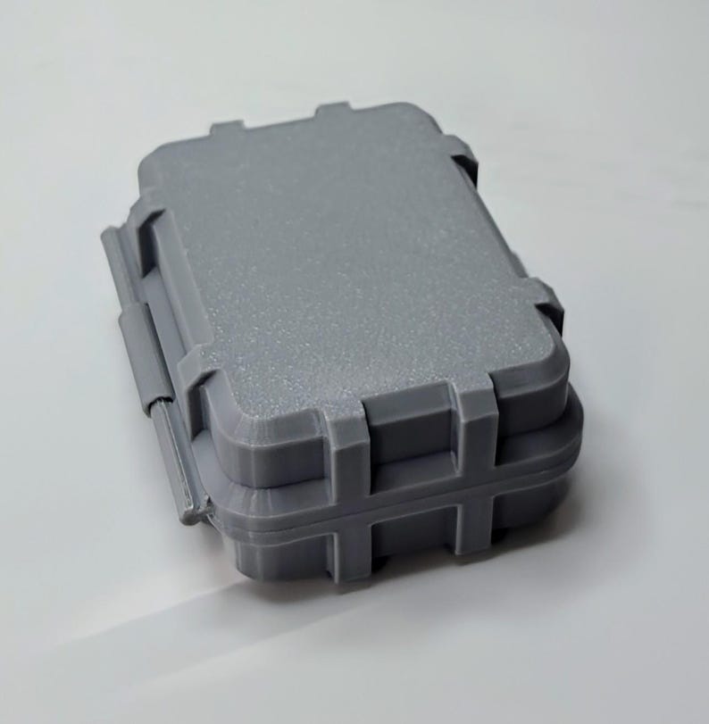May include: A small, gray, rectangular case with a textured surface. The case has a hinged lid and secure latches. The case is designed to protect its contents.