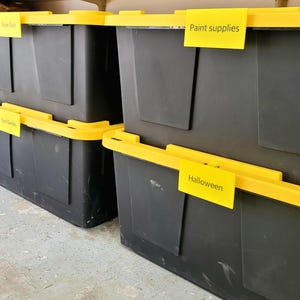 May include: Four black storage bins with yellow lids and labels. The labels read "Snow Gear", "Yard Games", "Paint Supplies", and "Halloween".