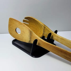 May include: Two wooden kitchen utensils, a spatula and a salad server, are resting in a black plastic stand. The utensils are made of natural bamboo wood.