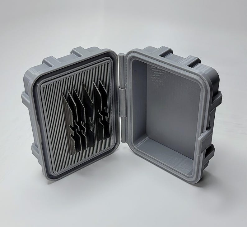 May include: Open gray plastic case with a ribbed interior and several dark gray, blade-like objects. The case is open, revealing the interior and the objects within. The case has a rectangular shape with a hinged lid.