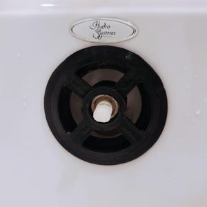 May include: Close-up of a black drain cover with a circular design, set in a white surface. The drain cover has a central white piece and is surrounded by a white surface. The text "Hydro Systems" is visible.