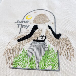 May include: Embroidered design on a cream-colored fabric featuring a stylized angel with tan wings and black arms. The angel wears a striped skirt and stands in front of green foliage. The text "June Tiny" is above the angel.