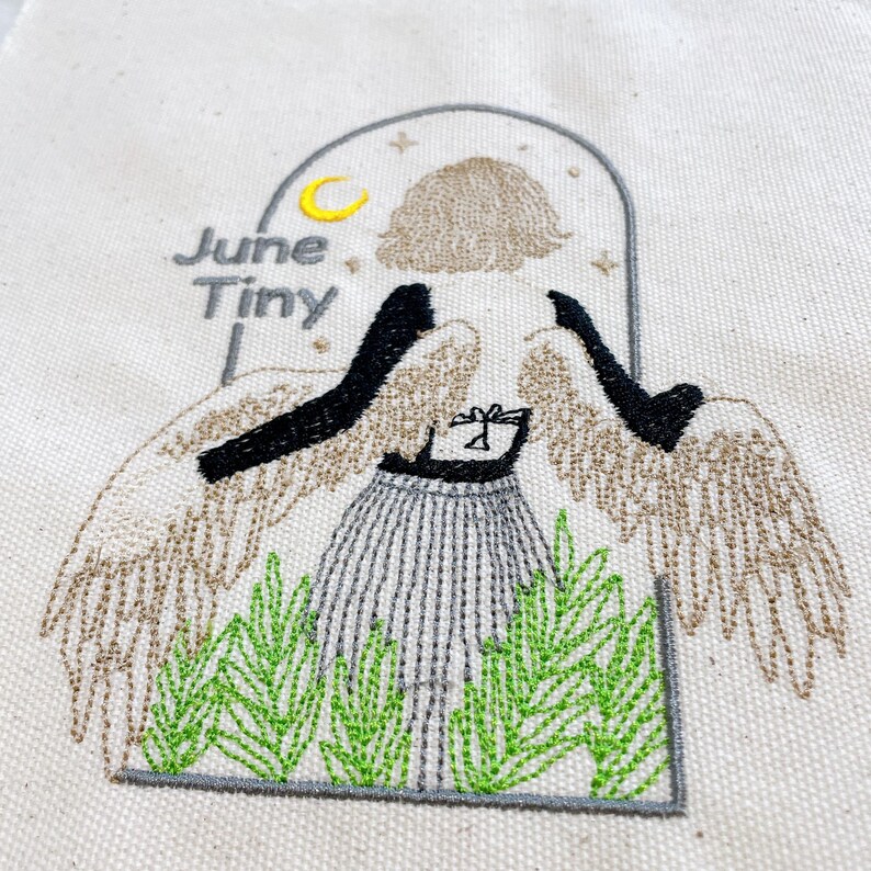 May include: Embroidered design on a cream-colored fabric featuring a stylized angel with tan wings and black arms. The angel wears a striped skirt and stands in front of green foliage. The text "June Tiny" is above the angel.