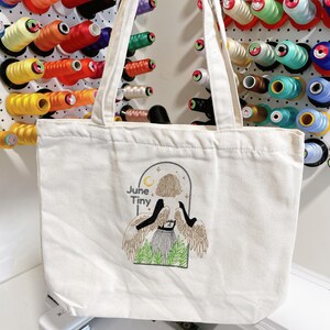 May include: Off-white canvas tote bag with embroidered design. The design features a figure with wings, a skirt, and the words "June Tiny". The bag has two handles and is displayed in front of colorful spools of thread.