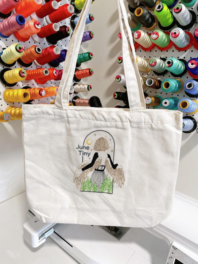 May include: Off-white canvas tote bag with embroidered design. The design features a figure with wings, a skirt, and the words "June Tiny". The bag has two handles and is displayed in front of colorful spools of thread.